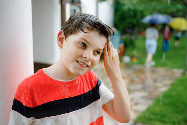 Школьник, бегущий сквозь сильный летний дождь в саду. Счастливый улыбающийся мокрый ребенок веселится с брызгами и прыжками в лужах. Мероприятия для детей в дождливую погоду. - Фото, изображение