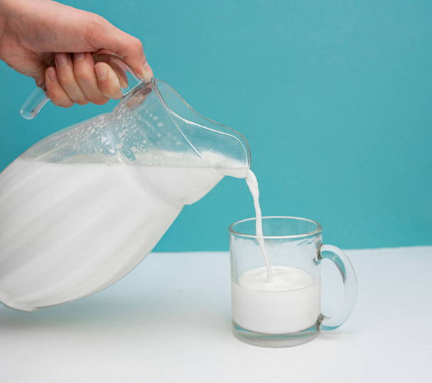 Fresh cow's milk is poured from a jug into a mug on a white and blue background. Milk Day. Healthy and tasty breakfast concept. copy space for text - Photo, image