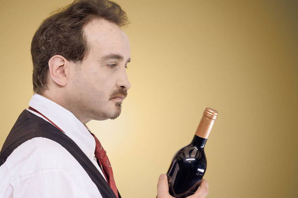 Uomo in camicia bianca, gilet e cravatta guardando una bottiglia di vino davanti a uno sfondo pastello chiaro - Foto, immagini