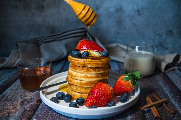 スティックから蜂蜜を注ぐ健康的なパンケーキの山,新鮮なブルーベリー,イチゴと木製のテーブルの上に牛乳のガラス,朝食のためのおいしいデザート - 写真・画像
