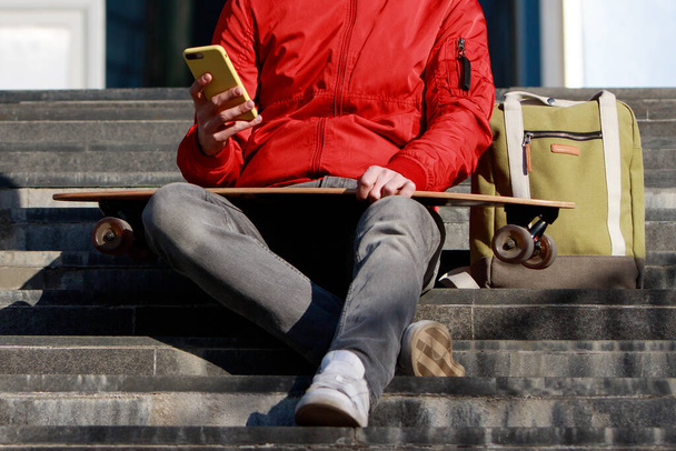 Stijlvolle longboarder in casual kleding met zijn smartphone, rustend op de treden, zittend met longboard / skateboard buiten, vooraanzicht, bijgesneden beeld. Urban, subcultuur, skateboarden concept - Foto, afbeelding
