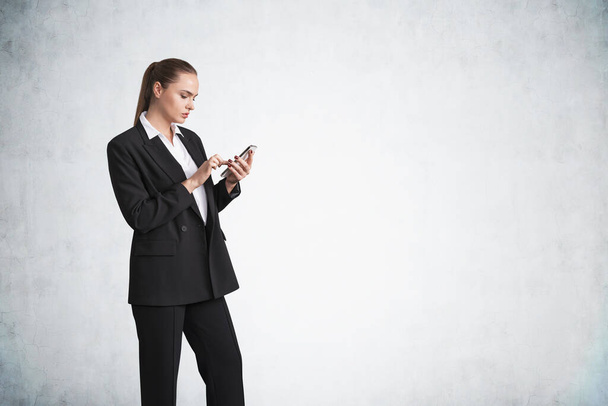 Office woman in black suit typing in phone. Pensive manager using mobile device. Mockup copy space blank wall, concept of business technology and app - Φωτογραφία, εικόνα