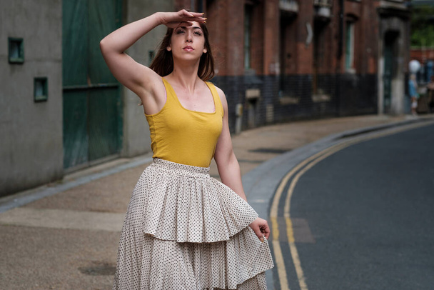 Mujer joven con una camiseta amarilla y falda blanca posando en una calle estrecha. Posturas de actuación y vibraciones malhumoradas. - Foto, imagen