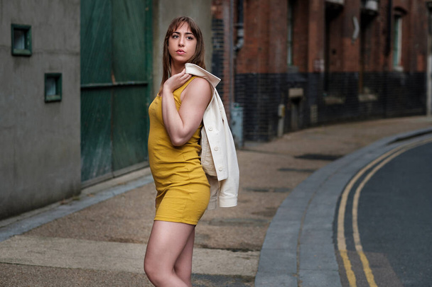 Młoda kobieta w żółtej sukience i białej kurtce pozująca na wąskiej ulicy. Moody sesji mody. - Zdjęcie, obraz
