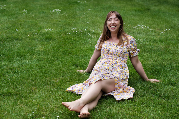 Entspannte junge Frau, die an einem Frühlingstag im Gras sitzt. Sie trägt ein Frühlingskleid. - Foto, Bild