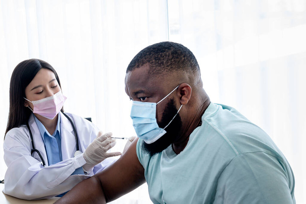Asian attractive woman doctor, wearing a surgical mask, is administering a vaccination treatment to an African American man patient, to health care and vaccine for people concept. - Photo, image