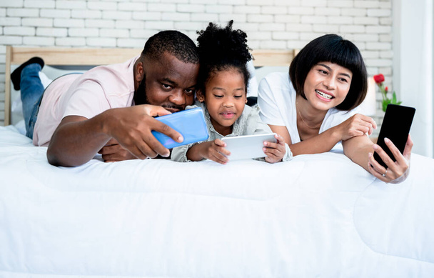 Családi program, afro-amerikai apa, egy ázsiai vonzó anya és egy 5 éves lány, aki egy fehér ágyban fekszik, és mobiltelefont használ a családi és technológiai koncepcióhoz.. - Fotó, kép