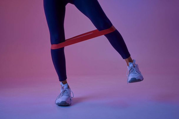Sexy female athlete doing exercise with rubber in studio, neon background. Fitness sportswoman at the photo shoot, sport concept, active lifestyle motivation - Foto, afbeelding
