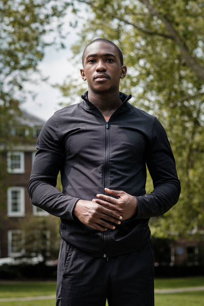 Retrato de un atleta negro posando en un parque. Está usando ropa negra activa.. - Foto, imagen