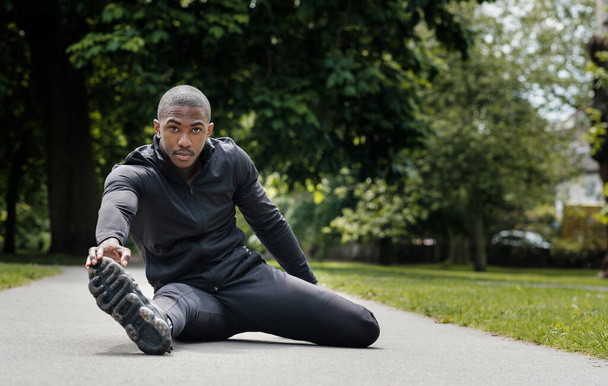 Retrato de un atleta negro que se extiende en un parque. Está usando ropa negra activa.. - Foto, Imagen