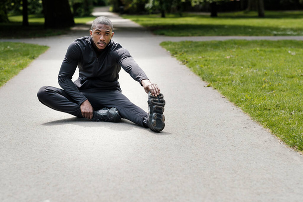 Portret van zwarte mannelijke atleet die zich uitstrekt in een park. Hij draagt zwarte actieve kleding.. - Foto, afbeelding