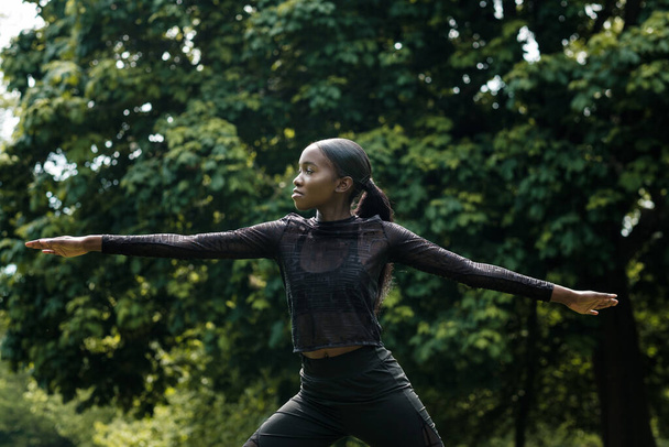 Portret van een zwarte vrouwelijke atleet die zich uitstrekt in een park. Ze draagt zwarte actieve kleding.. - Foto, afbeelding