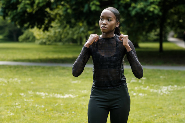 Porträt einer schwarzen Sportlerin, die sich in einem Park dehnt. Sie trägt schwarze Aktiv-Kleidung. - Foto, Bild