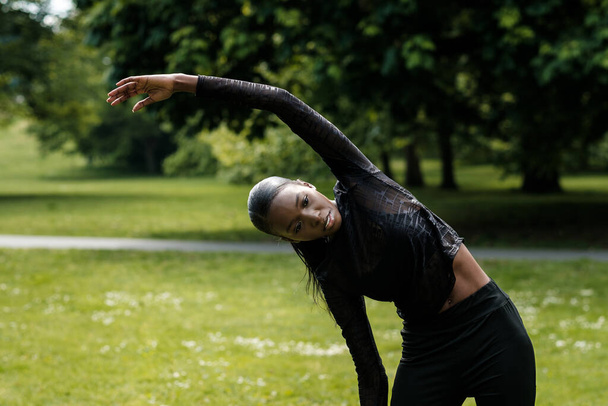 Портрет черной спортсменки, растянувшейся в парке. Она носит черную активную одежду. - Фото, изображение