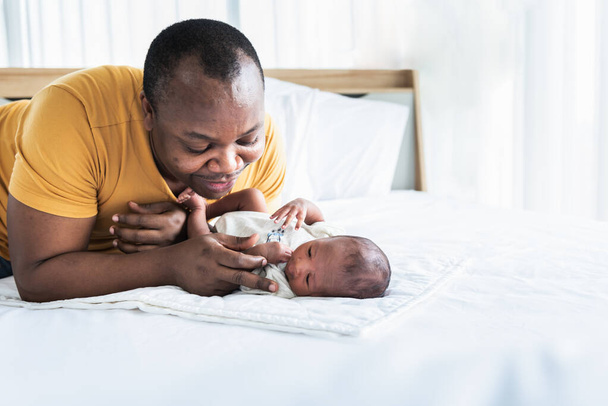 Afroamerykanin ojciec bawi się w flirt ze swoim 12-dniowym nowonarodzonym synem leżącym w łóżku w białej sypialni, ze szczęśliwą koncepcją dla afroamerykańskiej rodziny i noworodka - Zdjęcie, obraz