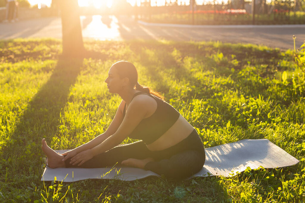 Tranquillo giovane donna incinta positiva in tuta da ginnastica fa yoga e meditare seduto su tappetino su erba verde nella calda giornata estiva soleggiata. Concetto di preparazione al parto e atteggiamento positivo - Foto, immagini