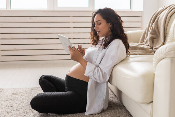 Pregnant woman holding digital while tablet sitting on a carpet near a couch in the living room at home - Photo, Image