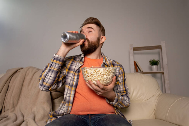 Bearded man watching film or sport games TV eating popcorn and drinking beer in house at night. Cinema, championship and fan concept. - Fotó, kép