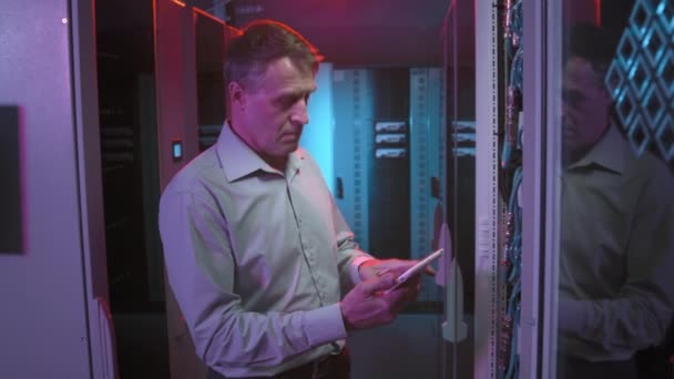 Imagem de rastreamento do técnico masculino de meia-idade digitando no tablet e verificando o equipamento de computação na sala de servidores do data center - Filmagem, Vídeo
