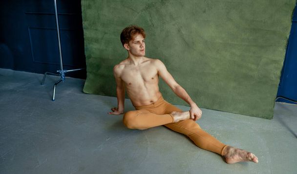 Male ballet dancer sittiing on the floor in dancing class, grunge wall on background. Performer with muscular body, grace and elegance of movements - Foto, imagen