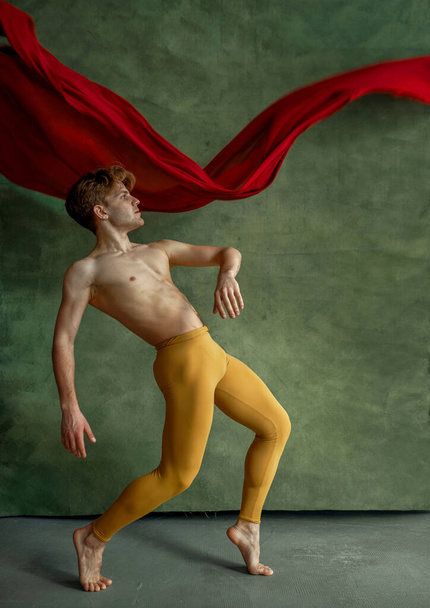Male ballet dancer doing exercise in dancing studio, grunge wall and red cloth on background. Performer with muscular body, grace and elegance of movements - Photo, Image