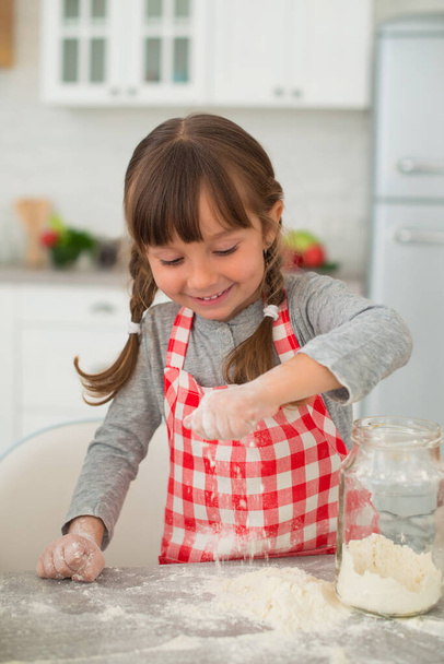 Cool smiling girl with pigtails in a checkered apron cooks in the kitchen, pours flour into the dough. - Foto, imagen