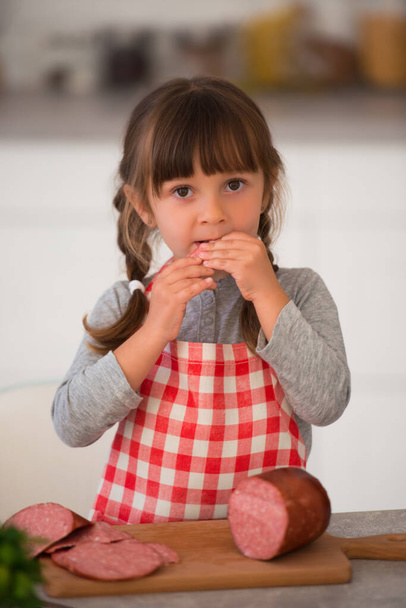 Cute little girl with pigtails in a checkered culinary apron eating sausage in the kitchen at the table. - Photo, image