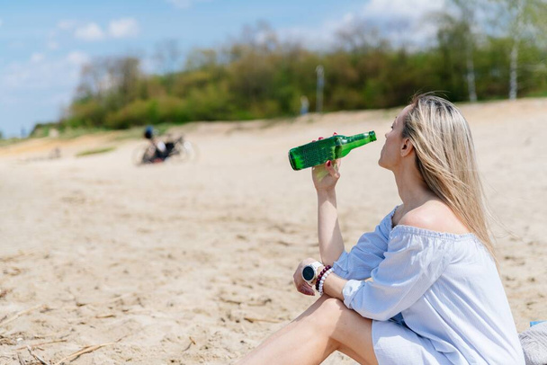 Atrakcyjna kobieta trzyma latem butelkę zimnego piwa na plaży nad jeziorem. - Zdjęcie, obraz