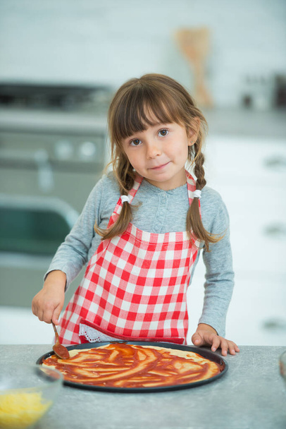 A funny little girl with pigtails in a checkered apron prepares pizza on her own in the kitchen. Cooking class for children. - Fotoğraf, Görsel