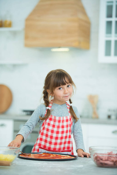 A funny little girl with pigtails in a checkered apron prepares pizza on her own in the kitchen. Cooking class for children. - Valokuva, kuva