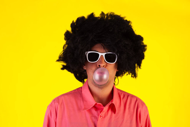Curly man wearing sunglasses blowing bubble with chewing gum on yellow background - Photo, Image