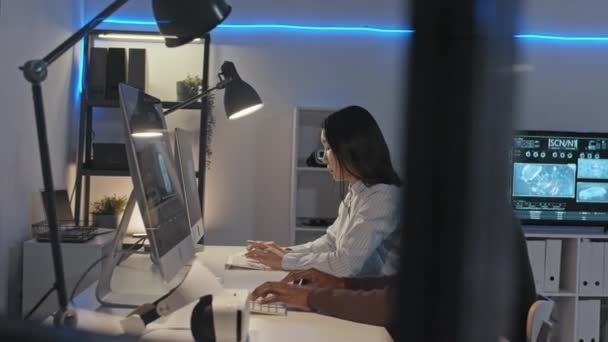 Tracking shot of male African-American and female Asian engineers sitting at desks in office and typing on keyboards Er is holografisch beeld van prototypes en gegevens op computerschermen - Video