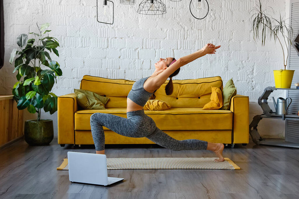 Fitness Online. Uma jovem atlética se estende na sala de estar, senta-se em um tapete de ioga na frente de um laptop, copia o espaço. Linda morena trabalhando em casa usando tutoriais em vídeo. - Foto, Imagem