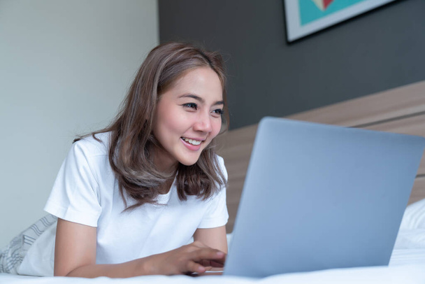 Gyönyörű ázsiai nő fekszik és mosolyog az ágyon és a laptopján. WFH Otthonról dolgozik a karanténban. Társadalmi elszigeteltség - Fotó, kép