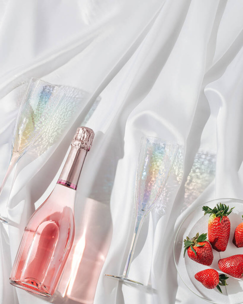 Rose champagne in bottle, bright glasses for wine and and red berries strawberry sparkling on light with beautiful shadows on white silk background. Celebration or relaxing evening. - Φωτογραφία, εικόνα