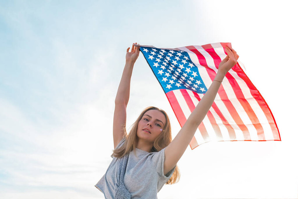 Młoda, tysiącletnia blondynka stojąca obok błękitnego nieba z amerykańską flagą. Flaga Stanów Zjednoczonych w jej rękach. Dzień Niepodległości 4 lipca. Święto narodowe USA. Koncepcja wolności i pamięci - Zdjęcie, obraz