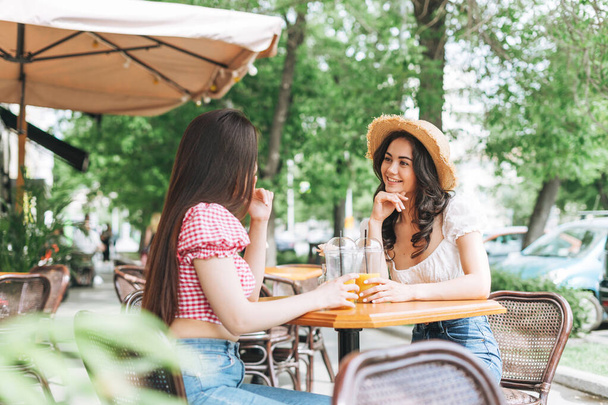 Happy smiling beautiful brunette young women friends in summer clothes with juice in hands sitting in cafe at summer city street - Foto, imagen