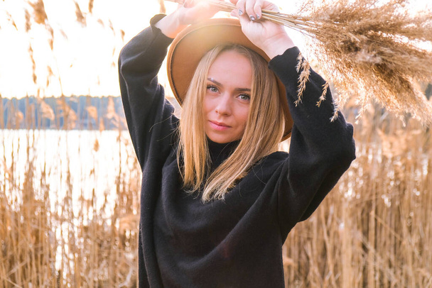 European blonde woman with beige hat in black sweater in the countryside. Golden hour, cottagecore. Local travel. Slow living. Mental health. Pampas grass - Photo, Image