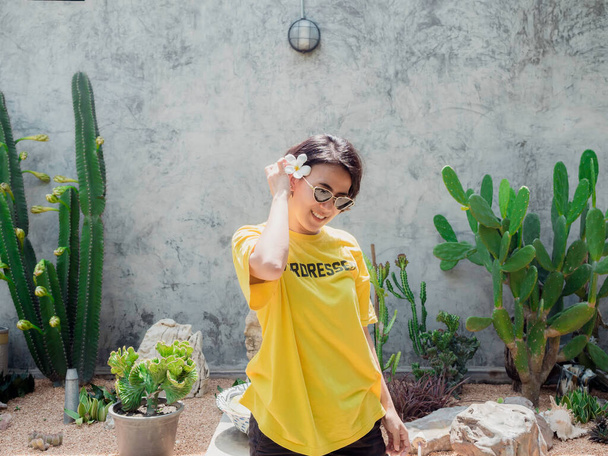 夏だ。太陽の光で笑顔。美しいアジアの女性の短い髪のサングラスと黄色のシャツを身に着けている彼女の耳の上に白いチュメリアの花を置くサボテンの庭と具体的な壁の背景. - 写真・画像