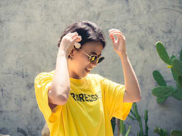 夏だ。太陽の光で笑顔。美しいアジアの女性の短い髪のサングラスと黄色のシャツを身に着けている彼女の耳の上に白いチュメリアの花を置くサボテンの庭と具体的な壁の背景. - 写真・画像