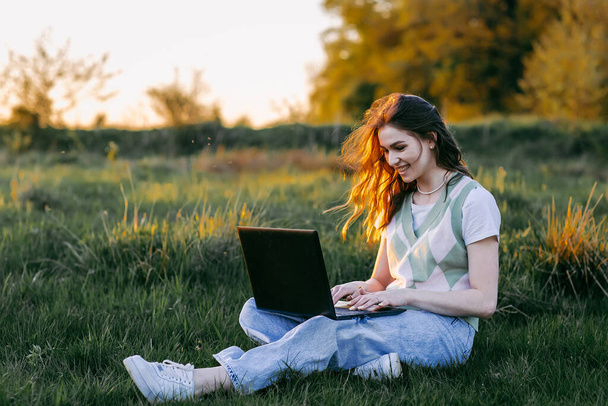 chica estudiante feliz trabajando con un ordenador portátil sentado en la hierba verde en el parque, la luz del atardecer. Concepto de estilo de vida, - Foto, imagen