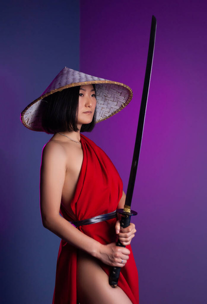 nackte schlanke Asiatin in rotem Umhang und asiatischem Hut mit einer Katana in der Hand Bild eines Samurai auf neonfarbenem Hintergrund - Foto, Bild