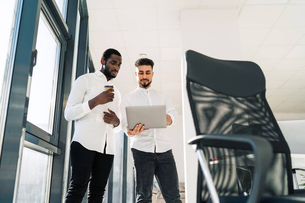 Два серьезных бизнесмена стоят в офисе с ноутбуком и обсуждают новый проект - Фото, изображение