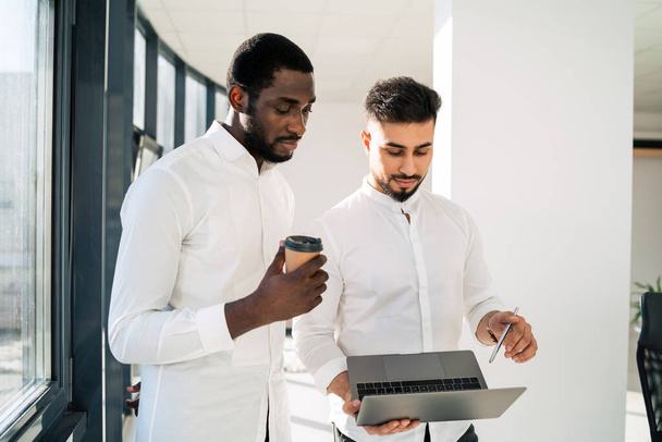 Dos jóvenes empresarios masculinos discutiendo la nueva startup mientras están de pie en medio de la oficina con el ordenador portátil - Foto, imagen