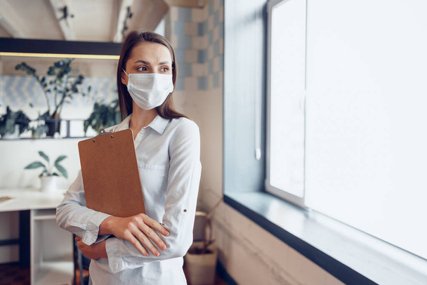 Young businesswoman in face mask standing in office and holding clipboard with documents - Fotó, kép