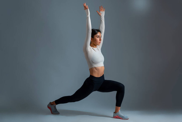 Woman in lunging Warrior Pose with arms outstretched up on grey background - Photo, Image