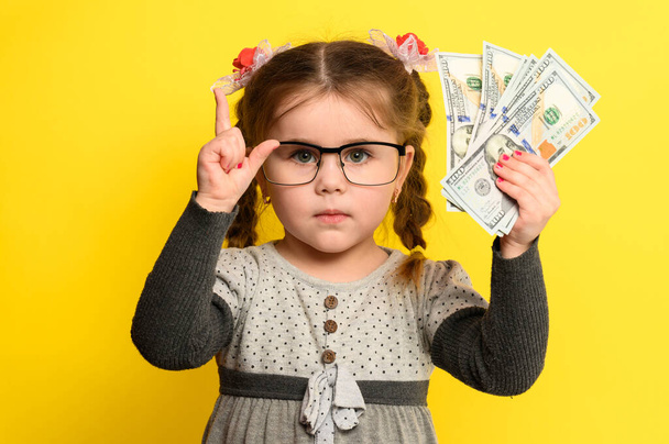 Dinheiro e crianças em segundo plano, uma menina rica com óculos segurando uma grande soma de dinheiro. novo - Foto, Imagem
