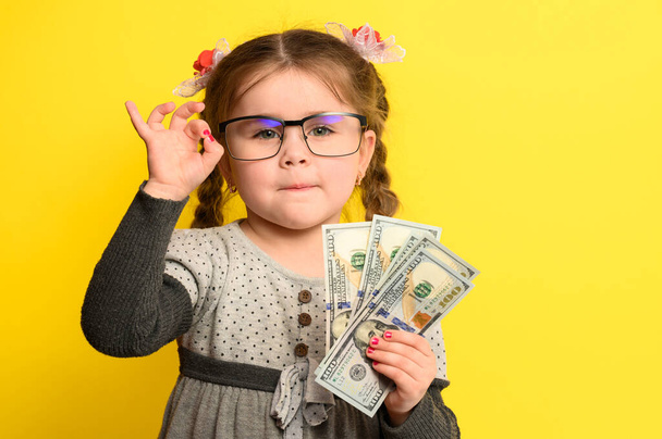 Geld und Kinder im Hintergrund, ein reiches Mädchen mit Brille, das eine große Summe Geld in der Hand hält. neu - Foto, Bild