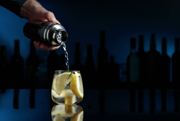 Koktajl "Gin-tonic" na czarnym tle odblaskowym. Barman nalewa koktajl z shakera do szklanki z lodem.. - Zdjęcie, obraz