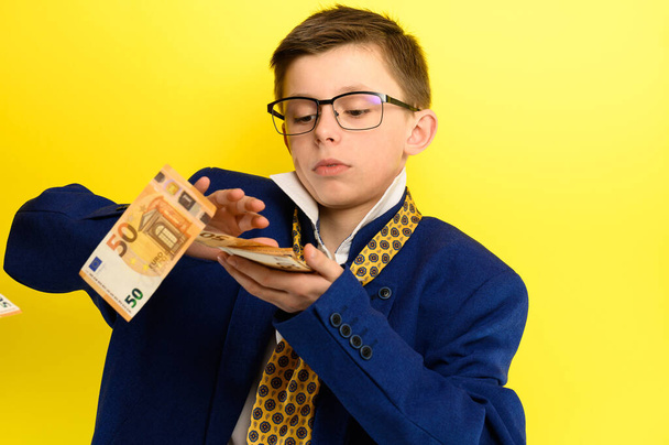 Dítě rozhazuje peníze, chlapec v obleku hází eura, portrét dítěte na žlutém pozadí. nový - Fotografie, Obrázek